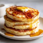 Close-up of a tall stack of fluffy buttermilk pancakes topped with melting butter and drizzled with syrup.