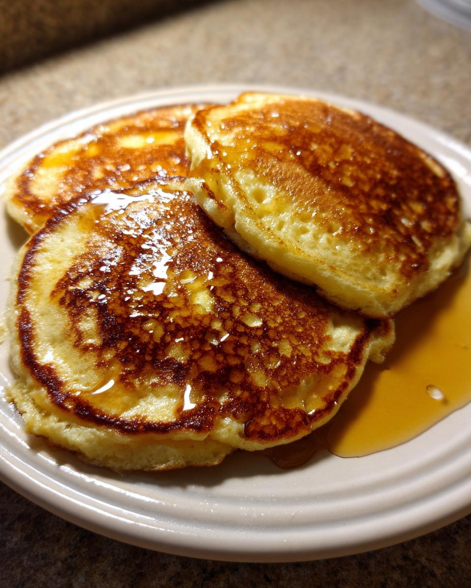 Three golden brown buttermilk pancakes stacked slightly, glistening with maple syrup.