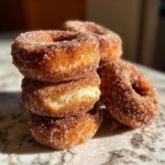 A stack of three fluffy, golden brown mini donuts coated heavily in cinnamon sugar, with one donut showing a soft interior.
