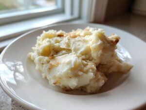 A serving of fluffy, creamy garlic mashed potatoes with slight golden-brown bits, presented on a white plate near a window.