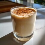 A close-up of a glass filled with creamy, frothy coquito recipe, dusted generously with ground cinnamon.