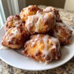 A close-up of several golden brown apple fritter bites drizzled with sweet white glaze, piled on a white plate.