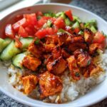 Close-up of a bowl featuring white rice topped with glazed, spicy harissa chicken pieces and fresh chopped tomatoes and cucumbers.
