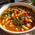 A close-up view of a white bowl filled with rich, tomato-based minestrone soup featuring elbow macaroni and wilted spinach.