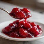 A spoonful of glistening, rich red homemade cherry pie filling dripping back onto a small white plate.