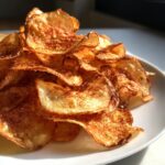 A close-up shot of a pile of golden, crispy homemade potato chips seasoned with paprika, served on a white plate.