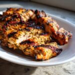 Close-up of perfectly grilled and sliced greek chicken pieces seasoned with herbs on a white plate.