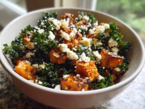 A close-up of a bowl containing a vibrant sweet potato salad mixed with dark green kale and topped with crumbled white feta cheese.