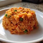 A molded serving of vibrant orange Mexican rice mixed with bright green peas and diced carrots on a white plate.