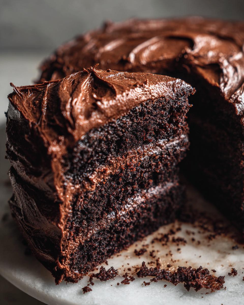 Close-up of a moist, dark slice of chocolate cake with thick, fudgy chocolate frosting, resting on a marble surface.