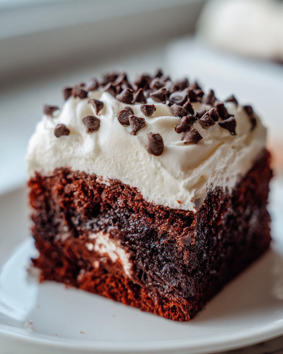 A close-up of a rich, moist slice of chocolate poke cake topped with white frosting and mini chocolate chips.