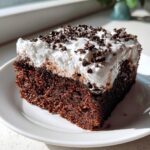 A close-up of a moist slice of chocolate poke cake topped with thick white frosting and chocolate shavings.