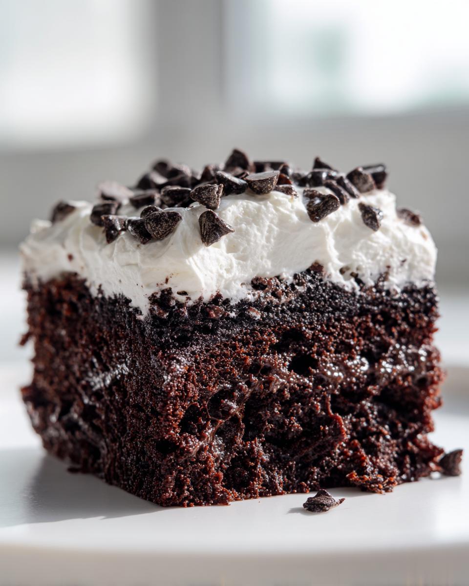 Close-up of a moist slice of chocolate poke cake topped with white whipped cream and mini chocolate chips.