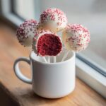 Four red velvet cake pops dipped in white chocolate and sprinkles, one is bitten showing the moist red interior.