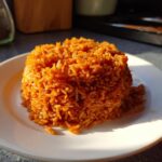 A mound of vibrant, reddish-orange Jollof rice served neatly on a white plate under warm sunlight.