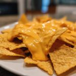 Close-up, appetizing shot of golden tortilla chips smothered in creamy, bright orange nacho cheese sauce.