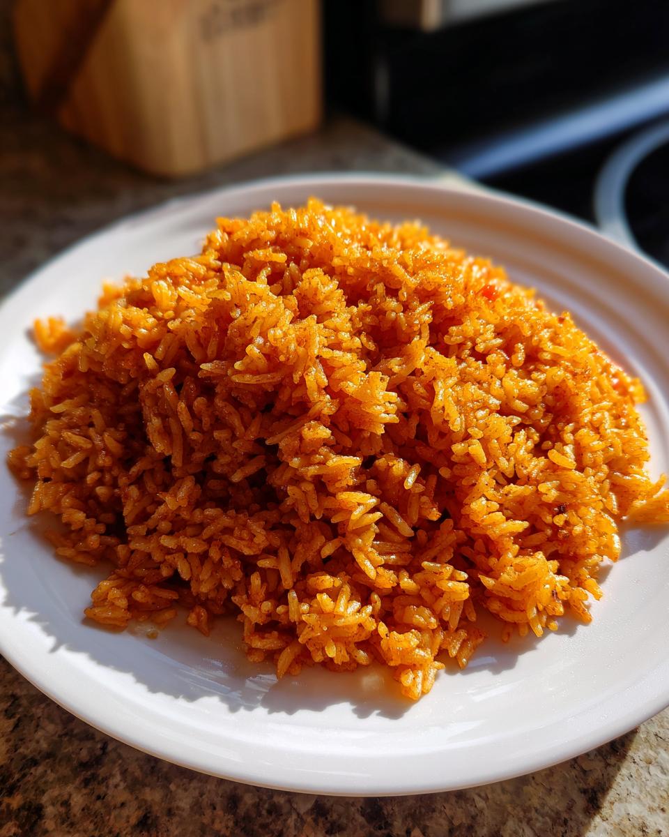 A mound of vibrant orange, fluffy jollof rice served on a white plate, glistening in the sunlight.