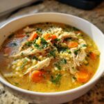 Close-up of a steaming bowl of penicillin soup featuring shredded chicken, bright orange carrots, rice, and fresh parsley garnish.
