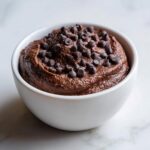 A close-up of thick, rich brownie batter dip served in a white bowl and generously topped with mini chocolate chips.