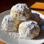 A close-up of four delicious russian tea cakes heavily dusted with powdered sugar on a white plate.
