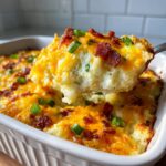 A spoonful of creamy mashed potato casserole topped with melted cheddar cheese, bacon bits, and green onions being lifted from the dish.