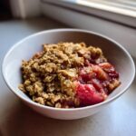Close-up of a single serving of warm rhubarb crisp with a thick, golden oat topping in a white bowl.