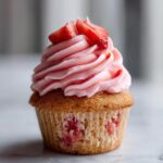 A close-up of a single, delicious strawberry cupcake topped with swirled pink frosting and fresh strawberry pieces.