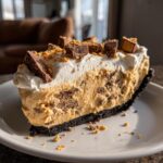 A rich slice of no-bake butterfinger pie on a white plate, topped with whipped cream and chopped candy pieces.