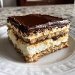 A square slice of layered eclair cake featuring graham crackers, white cream, and chocolate frosting.