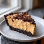 A decadent slice of peanut butter pie featuring a thick, creamy filling and a dark chocolate crumb crust, topped with chocolate crumbles.