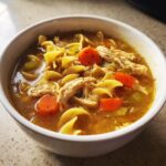 Close-up of a white bowl filled with rich broth, shredded chicken, egg noodles, and bright orange carrots from the slow cooker chicken noodle soup.
