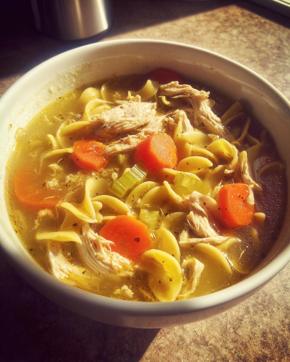 Close-up of a white bowl filled with rich broth, shredded chicken, egg noodles, carrots, and celery from slow cooker chicken noodle soup.