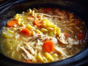 A close-up view of hot slow cooker chicken noodle soup featuring shredded chicken, bright orange carrots, and egg noodles in a savory broth.