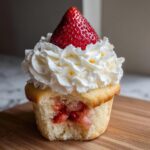 A close-up of a strawberry shortcake cupcake with a bite taken out, revealing strawberry filling, topped with whipped cream and a fresh strawberry.