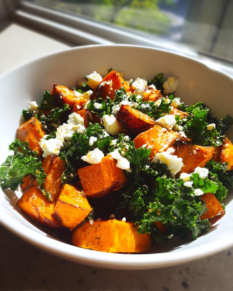 Close-up of a bowl of sweet potato salad featuring roasted orange sweet potato chunks, dark green kale, and crumbled white feta cheese.