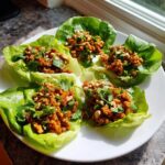 Four servings of flavorful thai chicken lettuce wraps, topped with chopped peanuts and cilantro, served on a white plate.