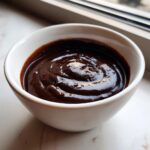 A close-up of thick, glossy homemade chocolate sauce recipe in a small white bowl.