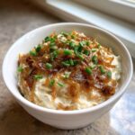 A white bowl filled with creamy caramelized onion dip, topped generously with dark caramelized onions and fresh green chives.