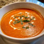 Close-up of a rich, orange tomato bisque garnished with a swirl of cream and fresh basil.