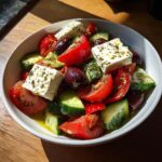 Close-up of a vibrant greek salad featuring chunks of tomato, cucumber, Kalamata olives, and large blocks of feta cheese drizzled with oil.