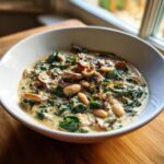 Close-up of a hearty white bean mushroom stew with spinach in a white bowl on a wooden surface.