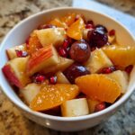 A close-up of a vibrant winter fruit salad featuring apples, oranges, grapes, and pomegranate seeds in a white bowl.