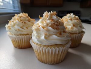 Three beautifully frosted coconut cupcakes topped with toasted coconut flakes sitting on a white counter.