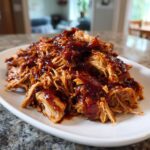 Close-up of shredded bbq chicken generously coated in dark, glossy barbecue sauce, served on a white platter.