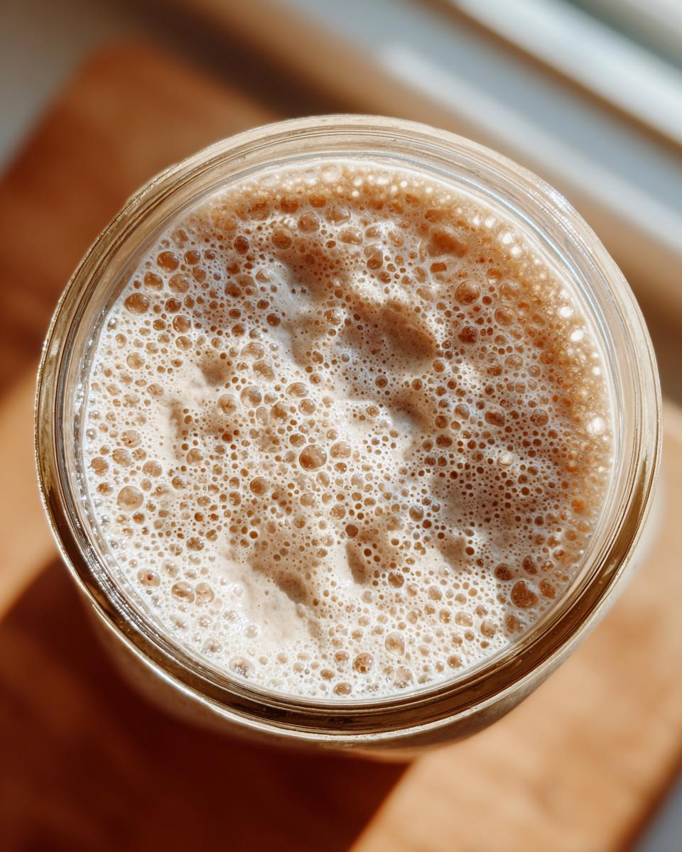 Overhead view of an active sourdough starter showing many small, vigorous bubbles in a glass jar.