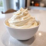 A close-up of fluffy, creamy white fruit dip swirled high in a small white bowl on a light countertop.