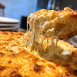A scoop of hot, gooey beer cheese dip being lifted, showing long, melted cheese strings stretching from the main dish.