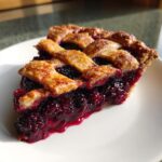 A close-up of a juicy slice of blackberry pie featuring a golden lattice crust and glistening, deep purple filling.