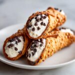 Three freshly filled cannoli pastries served on a white plate, topped with mini chocolate chips.