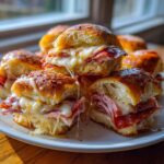A close-up of several hot grinder sliders stacked on a white plate, oozing with melted cheese and layered meats.